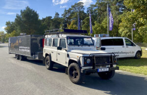 Weißer Land Rover Defender mit Ballonanhänger von aeroballonsport.de steht startklar als Rückholfahrzeug für eine Ballonfahrt in OWL auf einem Parkplatz vor Waldkulisse.