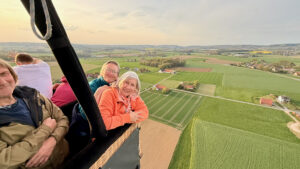 Gruppe lachender Passagiere lehnt am Korb eines Heißluftballons während einer Ballonfahrt über Kirchlengern und Herford in Ostwestfalen-Lippe, Blick auf Felder und Höfe in OWL.