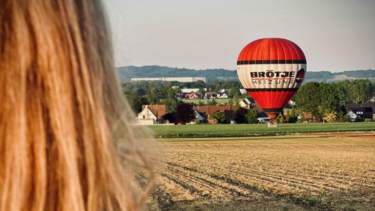 Person blickt über ein Feld auf einen rot-weißen Heißluftballon, der vor einer Ortschaft nahe Melle in Ostwestfalen-Lippe zur Landung ansetzt, Ballonfahrt in OWL NRW.