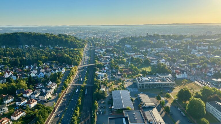 Panoramaaufnahme aus einem Heißluftballon über Bielefeld in der Abendsonne: Eine mehrspurige Straße und Bahntrasse verlaufen durch die Stadt, umgeben von Wohnhäusern, grünen Hügeln und modernen Gebäuden, im Hintergrund erstreckt sich die weite Stadtlandschaft bis zum Horizont.
