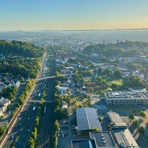 Panoramaaufnahme aus einem Heißluftballon über Bielefeld in der Abendsonne: Eine mehrspurige Straße und Bahntrasse verlaufen durch die Stadt, umgeben von Wohnhäusern, grünen Hügeln und modernen Gebäuden, im Hintergrund erstreckt sich die weite Stadtlandschaft bis zum Horizont.
