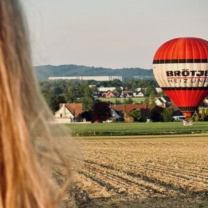 Person blickt über ein Feld auf einen rot-weißen Heißluftballon, der vor einer Ortschaft nahe Melle in Ostwestfalen-Lippe zur Landung ansetzt, Ballonfahrt in OWL NRW.